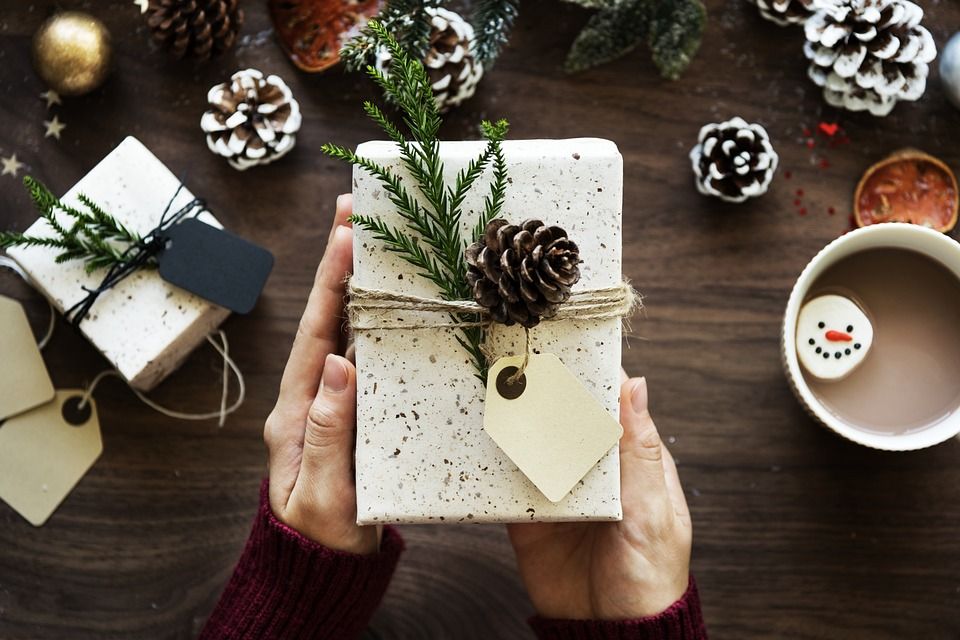 Tangan memegang paket hadiah Natal dengan tali dan label, ditemani oleh permen salju dan kopi hangat.
