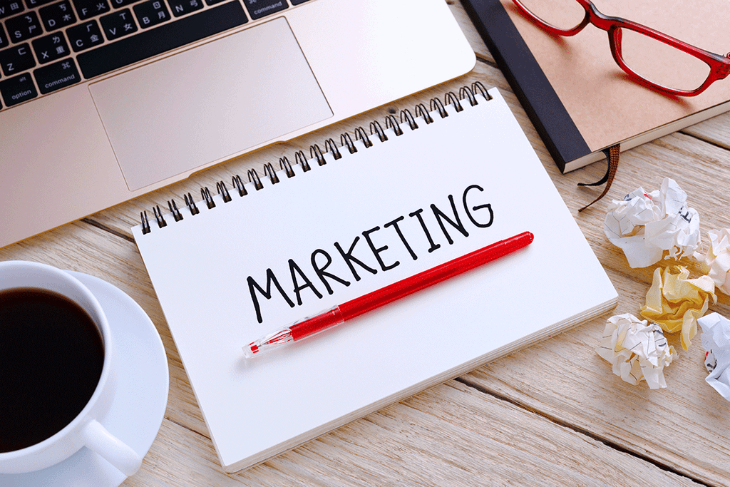 Notebook dengan tulisan 'MARKETING' dan pena merah, di samping laptop dan kaca mata.