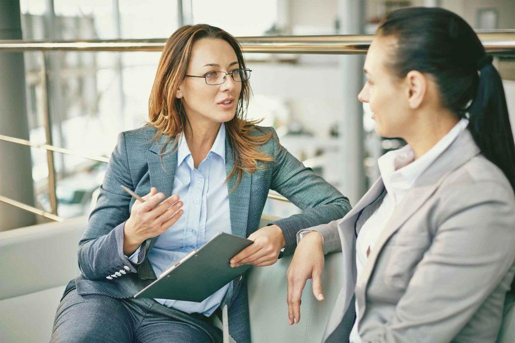 Dua wanita berbincang sambil menunjukkan dokumen di kantor modern.