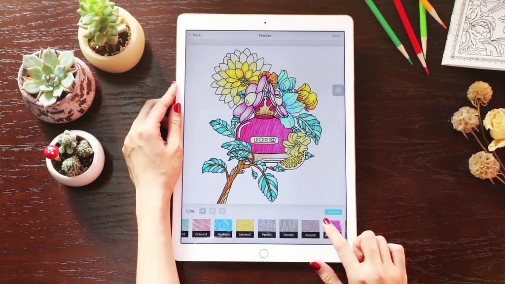 Tangan memegang tablet dengan aplikasi mewarnai dan potongan bunga di meja.