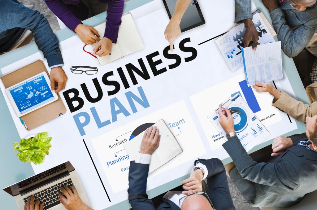 Grafik dan tangan menulis 'Business Plan' di meja pertemuan.