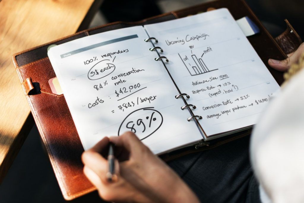 Tangan menulis analisis campaign di agenda dengan grafik dan angka.