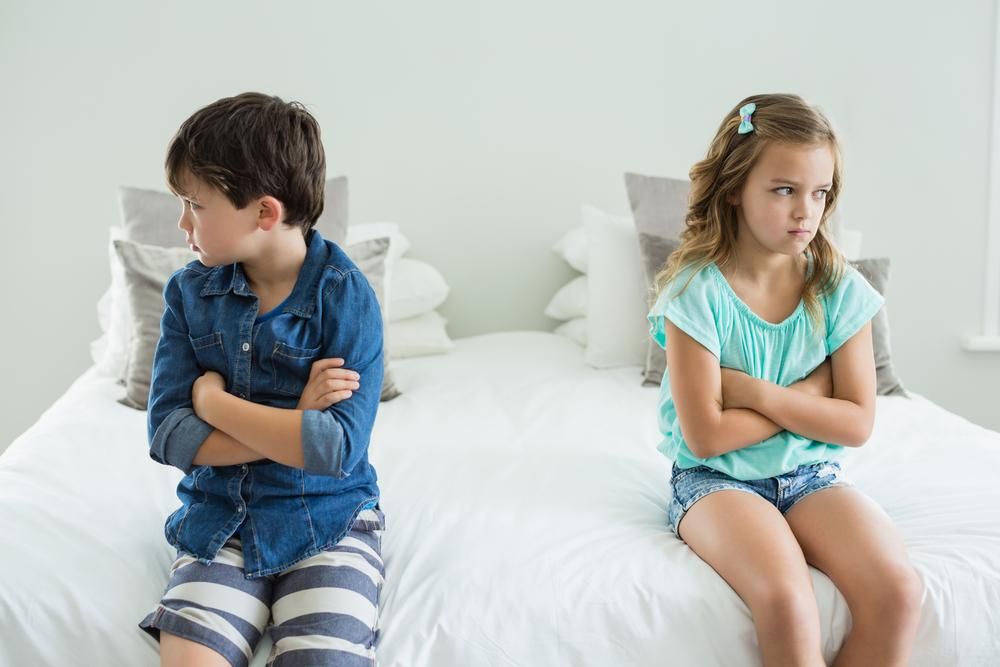 Dua anak kecil duduk di tempat tidur dengan tangan terlipat, tampak sedih.