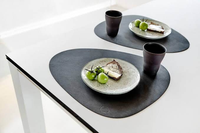 Tampilan peralatan makan modern dengan placemat hitam dan piring putih di atas meja putih.