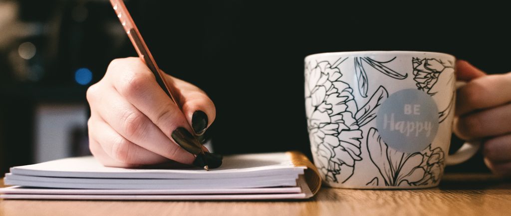 Tangan menggambar di buku dengan pena, sambil menengok mug motif bunga 'Be Happy'.