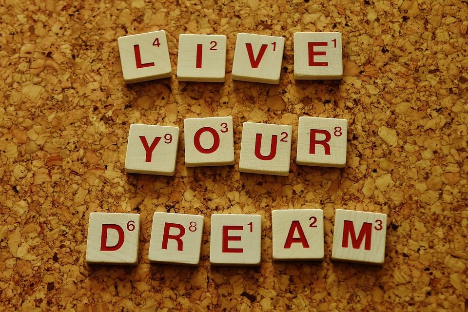 Gambar menunjukkan huruf Scrabble yang membentuk frasa 'Live Your Dream' di atas permukaan kayu.