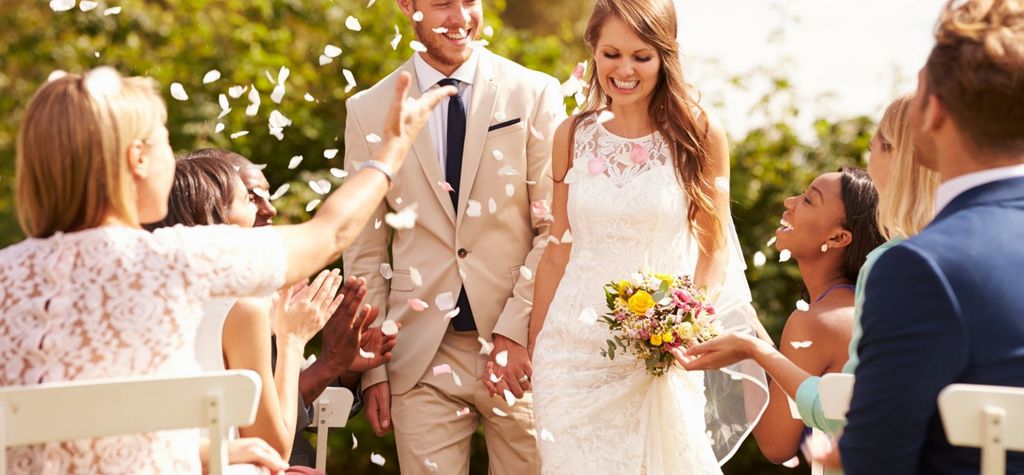 Pasangan pengantin berjalan di latar belakang taman hijau, diselubungi petal bunga putih.