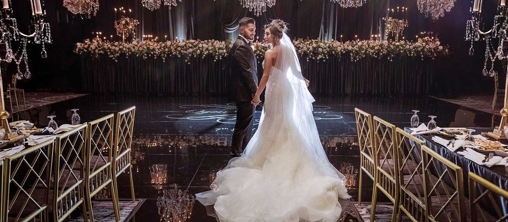 Pasangan pengantin menari di ruang mewah dengan dekorasi bunga dan lampu gantung.