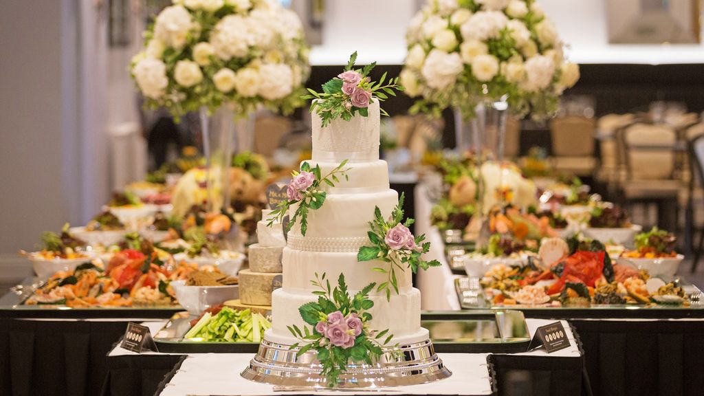 Kue pernikahan putih elegan dengan bunga mawar dan daun hijau, di depan meja hidangan.