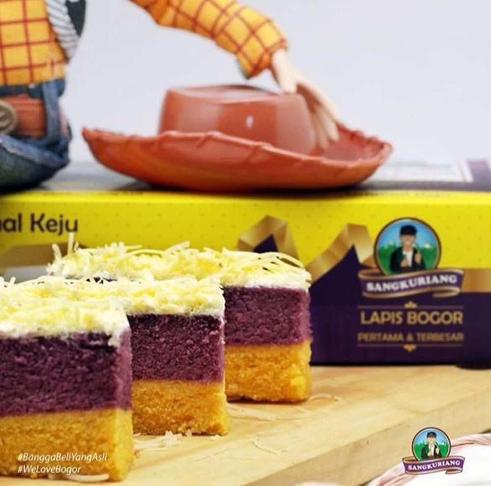 Kue lapis bogor keju dengan lapisan ungu dan kuning, dipotong dan disajikan dengan hati cowboy di atas kotak kemasan.