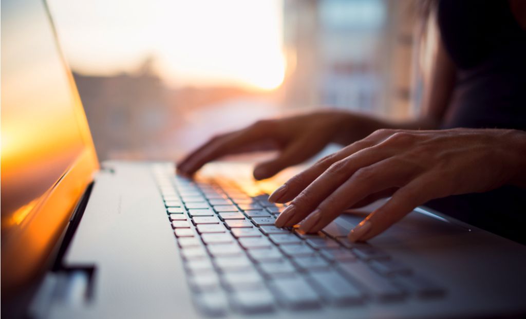 Tangan sedang mengetik pada keyboard laptop dengan cahaya matahari terbit di latar belakang.