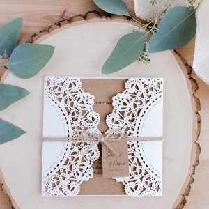Undangan elegan dengan doily putih dan tali di atas meja kayu dengan daun eucalyptus.