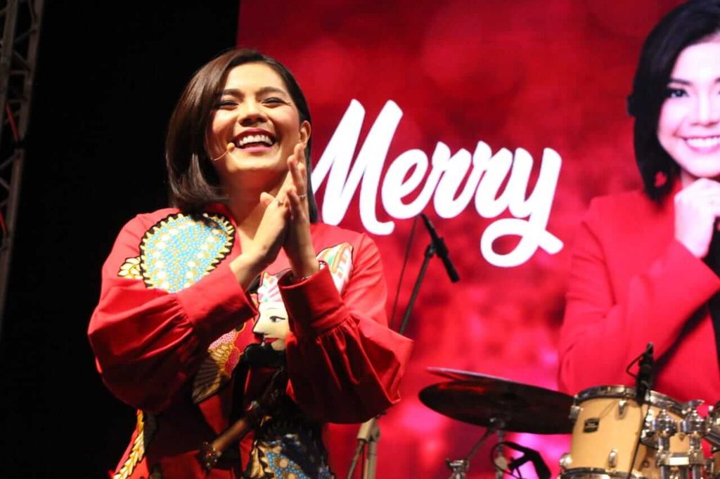 Seorang wanita sedang berterima kasih di depan latar Merry dan alat musik drum.