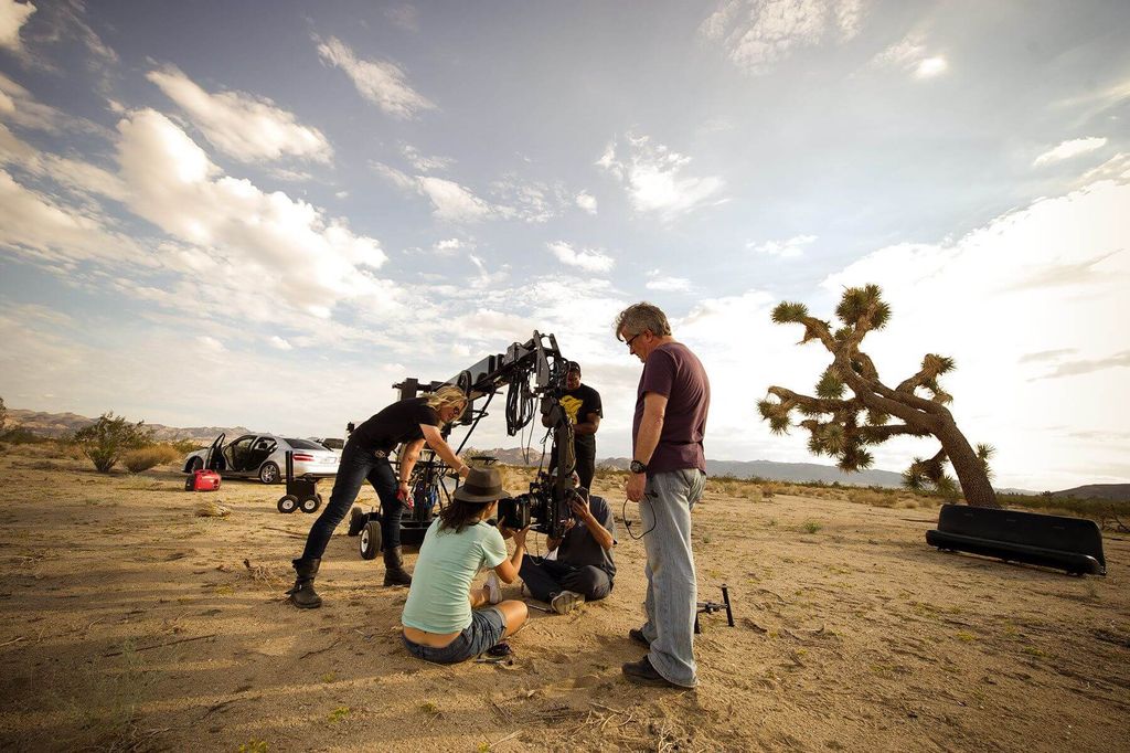 Tim produksi film sedang mempersiapkan peralatan di tengah padang pasir dengan pohon Joshua dan mobil di latar belakang.