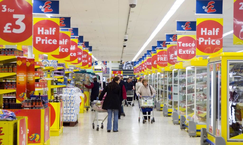 Interior of a supermarket aisle with promotional signs and shoppers pushing carts.