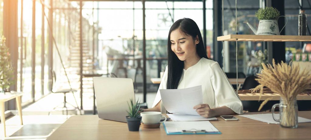 Perempuan memeriksa dokumen di depan laptop di cafe modern.