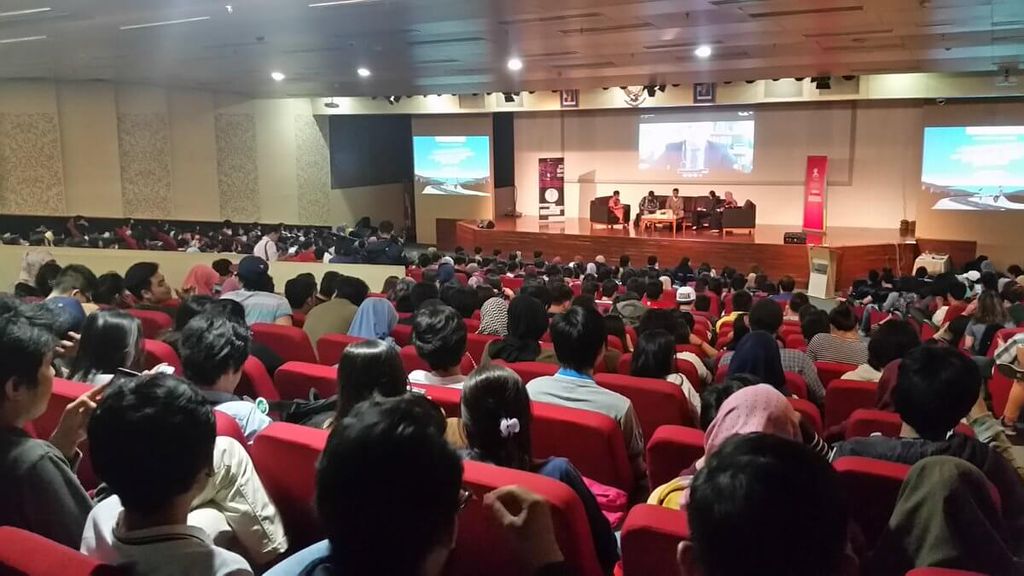 Pemandangan auditorium penuh penonton dengan layar presentasi dan panggung di belakang.