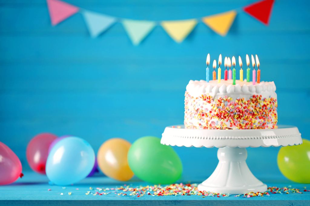 Kue ulang tahun berwarna cerah dengan lilin berwarna pastel dan balon di sekitarnya.