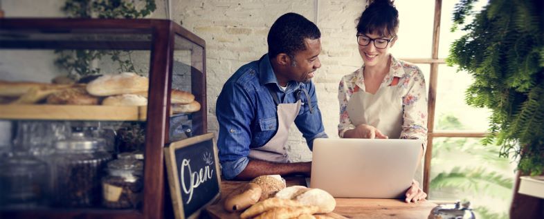 Dua orang pekerja berdiskusi di depan laptop di toko roti, dengan rak roti dan tanda 'Open' di latar belakang.