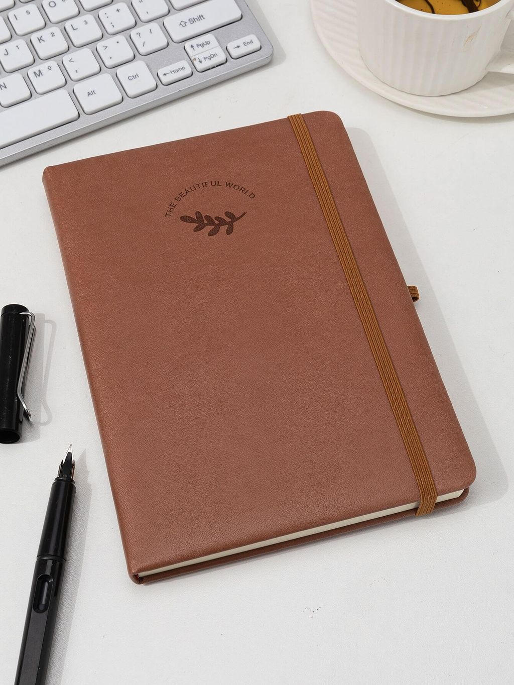 Notebook coklat dengan sablon 'The Beautiful World' dan tali elastis, ditempatkan di meja kerja.