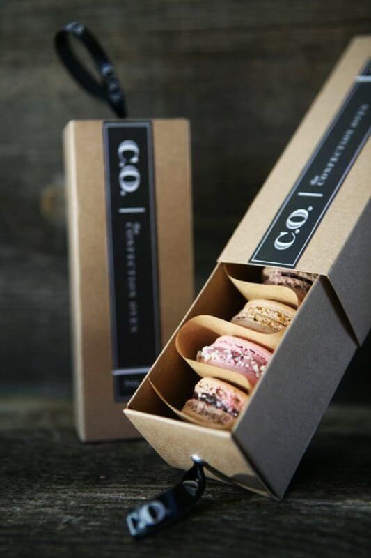 Kotak Macaron C.O. terbuka menampilkan berbagai warna macaron di atas meja kayu.