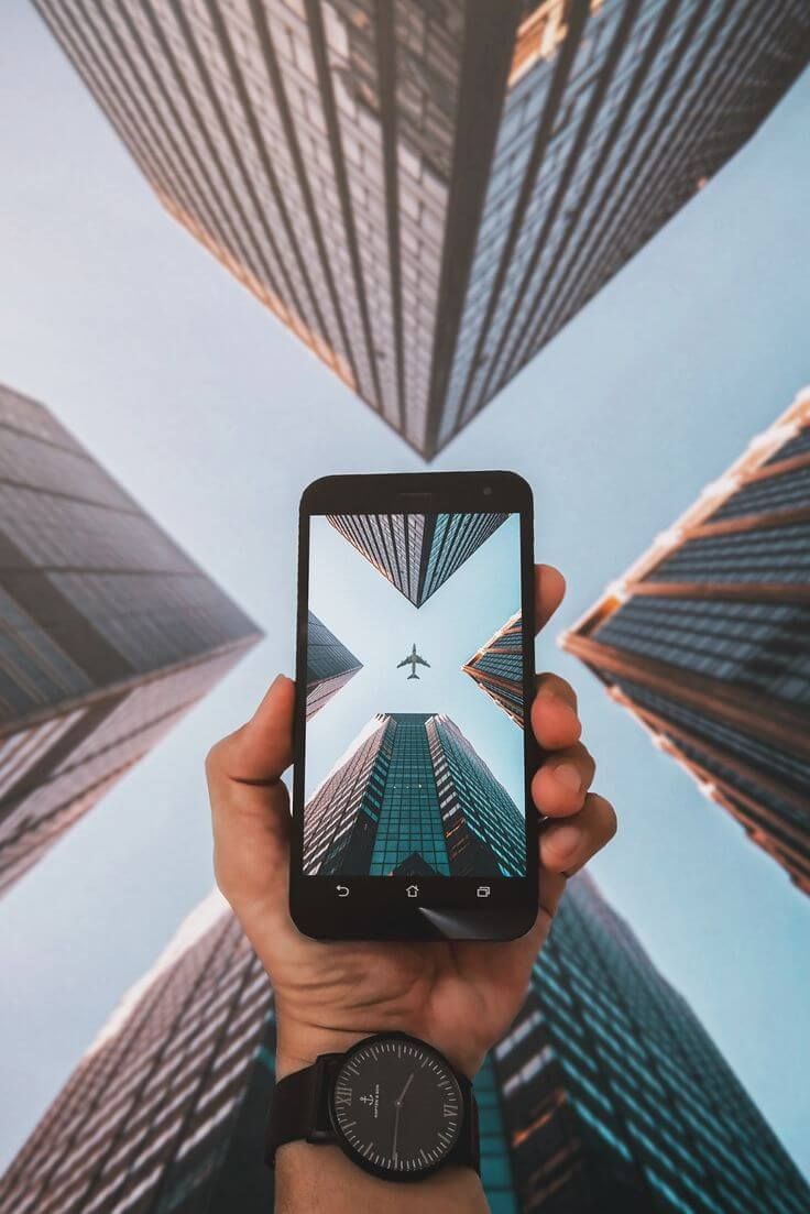 Tangan memegang smartphone menampilkan foto kota modern dengan bangunan tinggi dan pesawat terbang.