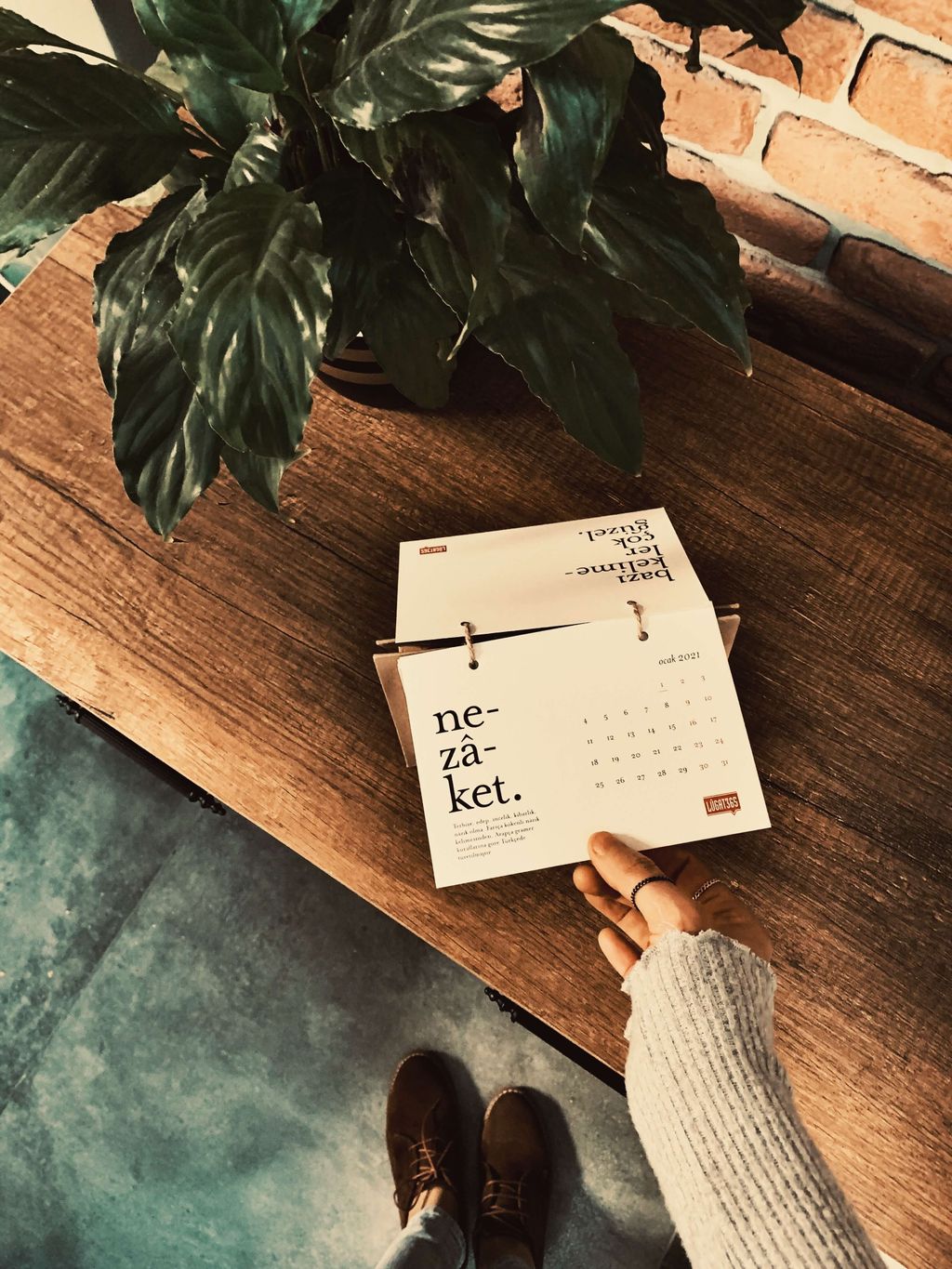 Tangan memegang kalender putih dengan tulisan 'ne-zá-ket' di meja kayu dengan tanaman dekoratif di belakang.