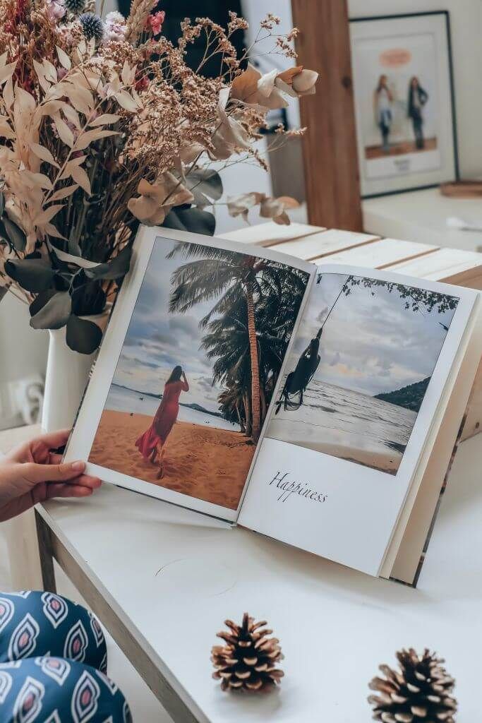 Tangan memegang buku foto terbuka dengan pemandangan pantai dan tulisan 'Happiness'.