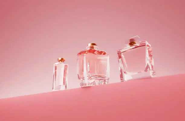 Tiga botol parfum berwarna transparan dengan tutup emas di latar belakang pink.
