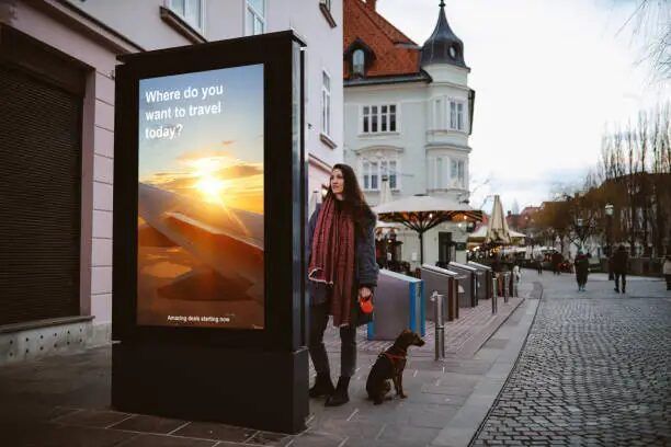 Perempuan berjalan di jalan kota dengan iklan digital di sebelahnya.
