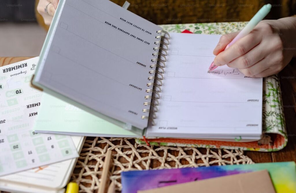 Tangan memegang pena dan menulis di buku agenda spiral.