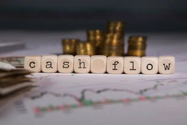 Tulisan 'cash flow' dari kubus kayu dengan latar belakang grafik keuangan dan koin.