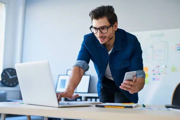 Desainer sedang bekerja di kantor dengan laptop dan smartphone.
