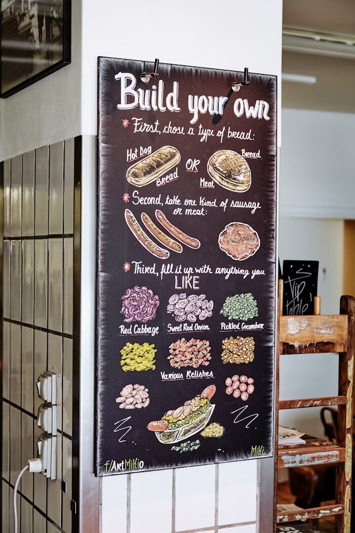 Papan menu untuk membuat sendiri hot dog dengan berbagai opsi bahan dan saus sebagai contoh materi promosi dekat titik keputusan beli di kafe.