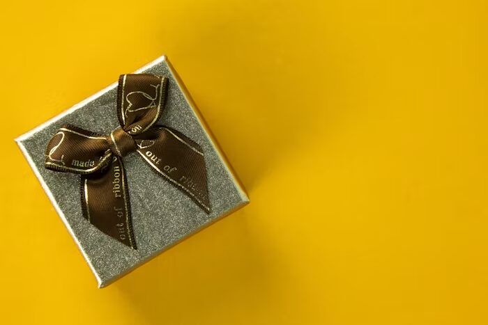Kotak hadiah kecil dengan pita emas di atas latar kuning cerah.