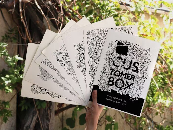Tangan memegang buku mewarnai dengan desain floral dan geometris, latar belakang tumbuhan hijau.