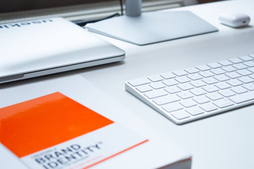 Meja kerja minimalis dengan laptop, keyboard, mouse, dan buku 'Brand Identity'.