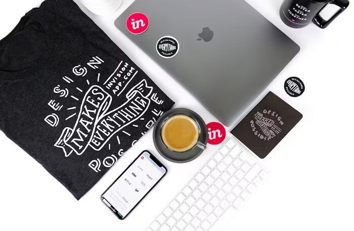 T-shirt, laptop, smartphone, dan aksesori percetakan online di meja kerja.