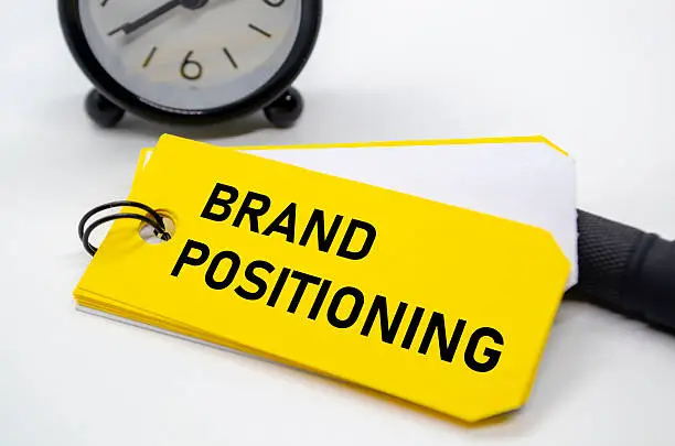 Label kuning dengan tulisan BRAND POSITIONING dan alarm di belakang sebagai ilustrasi pentingnya menentukan posisi merek