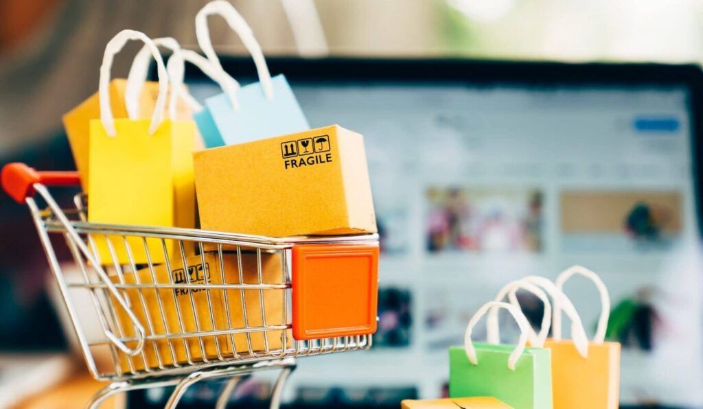 Mini keranjang belanja online berisi tas dan kotak kardus, layar laptop di latar belakang.