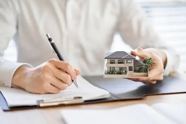 Tangan menulis dokumen dan memegang model rumah kecil, terkait analisis properti.