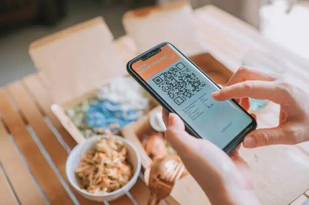 Tangan memegang smartphone dengan kode QR di layar, di depan makanan.