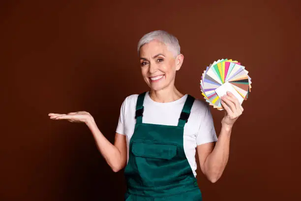 Perempuan memegang fan warna dan menunjukkan tangan kosong, latar coklat.