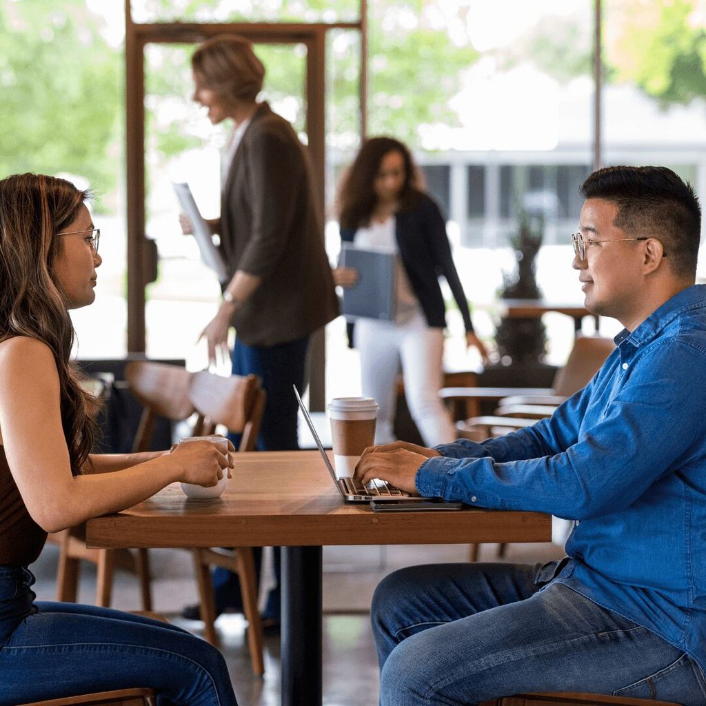 Dua orang sedang berbicara di meja kafe dengan laptop dan kopi.