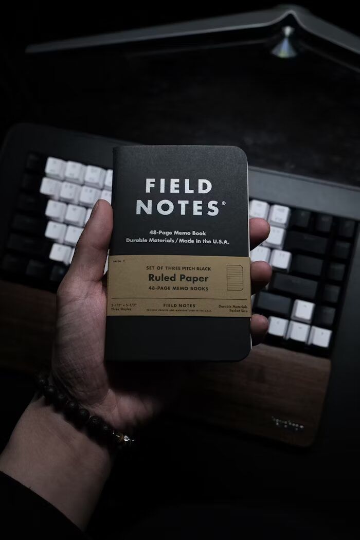Tangan memegang buku catatan Field Notes dengan latar keyboard di baliknya.