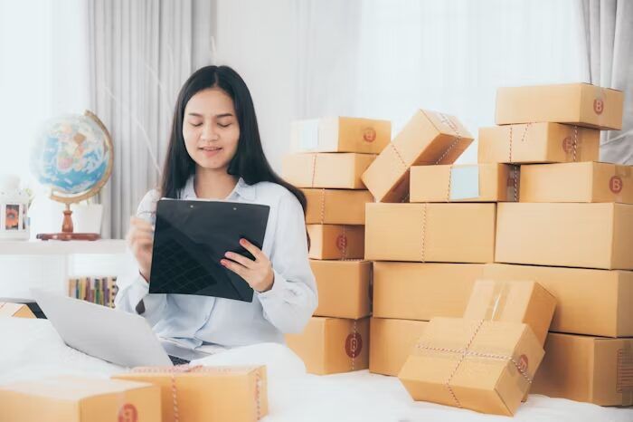 Perempuan sedang mengecek orderan di Uprint.id dengan tablet di sekitar kotak-kotak kemasan.