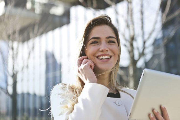Perempuan cantik tersenyum sambil memegang tablet di latar bangunan modern.