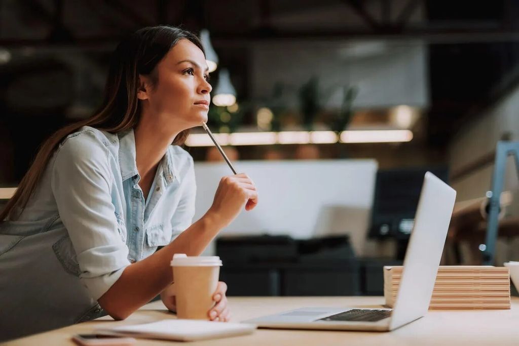 Wanita sedang bekerja di meja dengan laptop dan kopi, latar kantor modern.