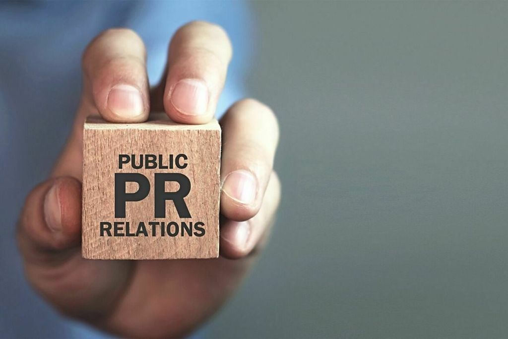 Tangan memegang blok kayu bertuliskan 'Public Relations'