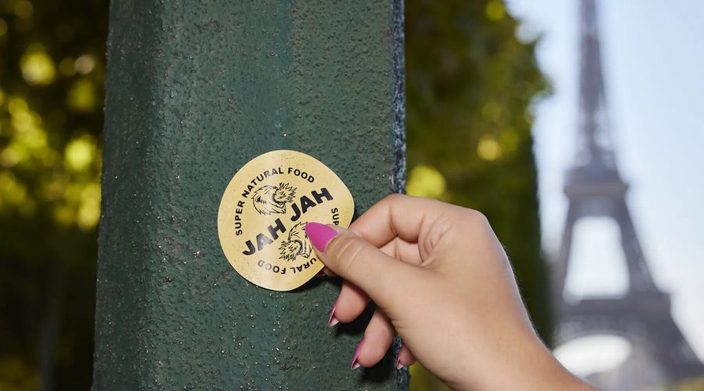 Tangan memegang stiker 'JAH JAH' dengan latar Menara Eiffel